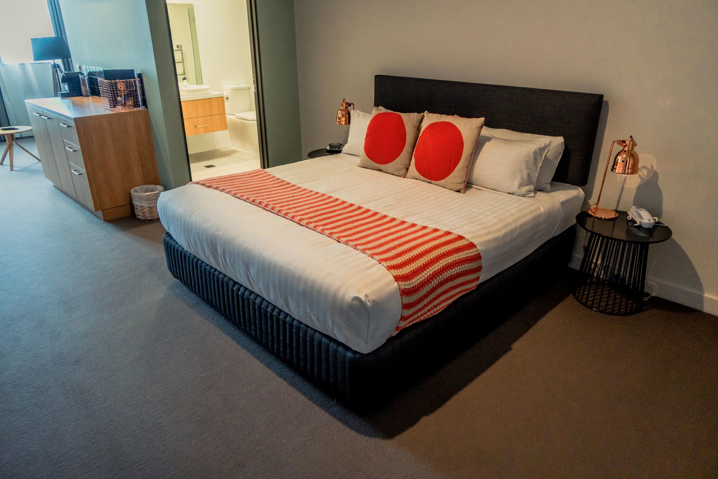 Een netjes opgemaakt bed met een gestreept dekbed, twee kussens met rode accenten, en een donker hoofdbord. Er staat een klein nachtkastje met een lamp en een telefoon.