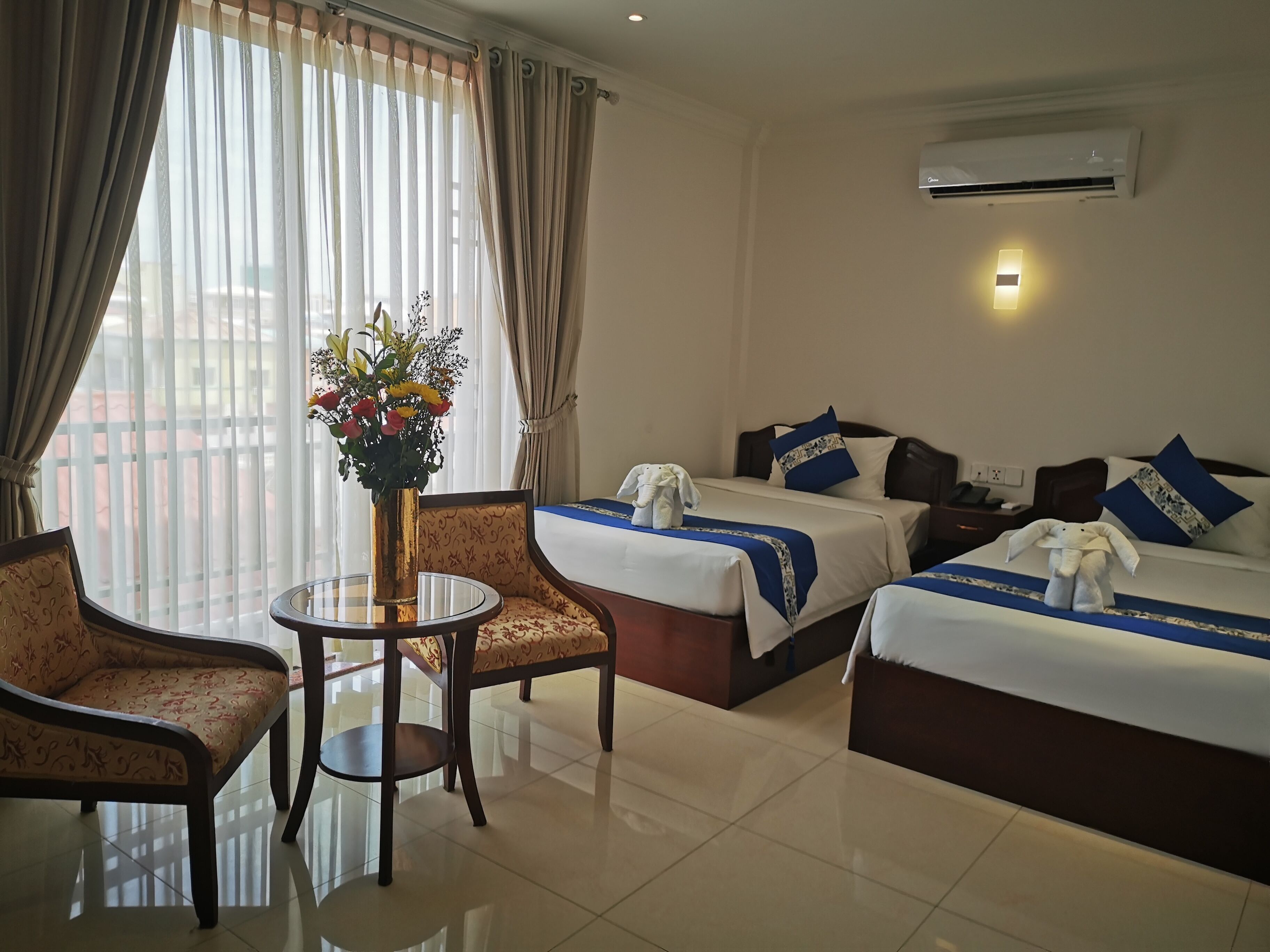 Habitación de hotel con dos camas, una zona de estar con mesa y sillas, y un ventanal con cortinas.