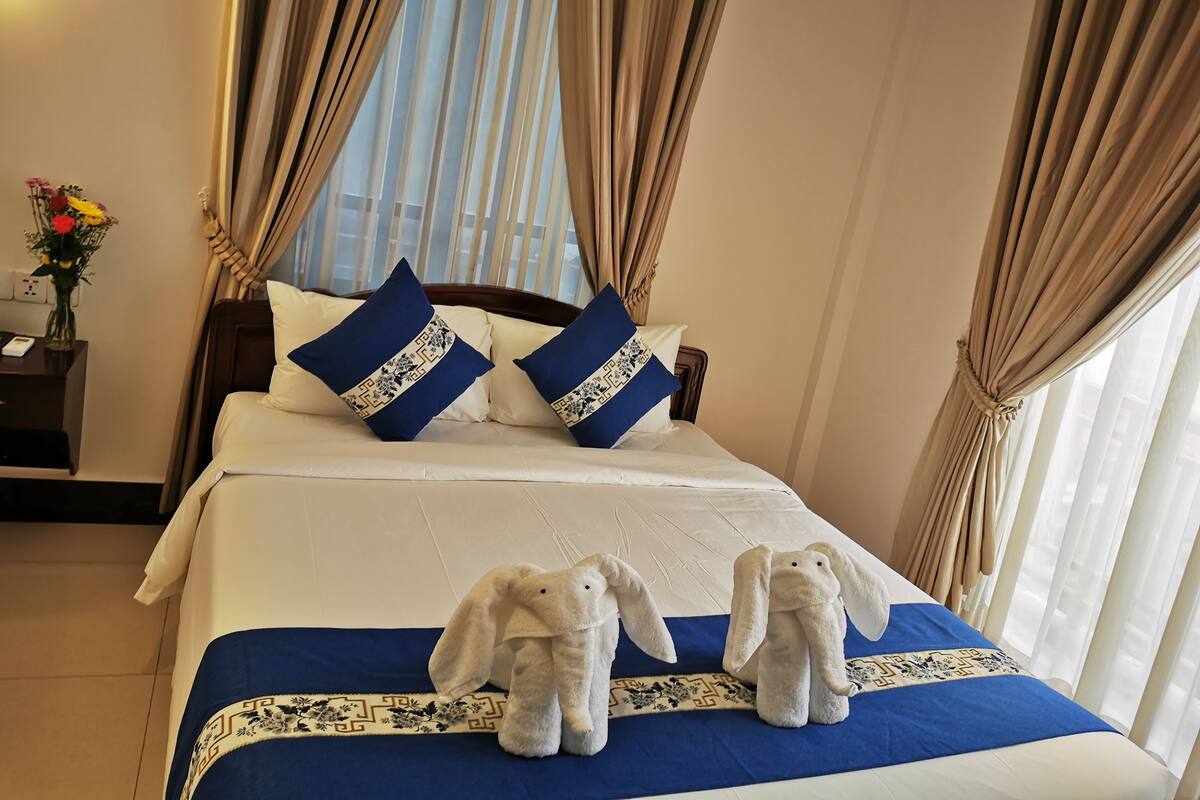 A neatly made bed with a blue bedspread and decorative pillows, a bedside table with a vase of flowers, and a view through sheer curtains.