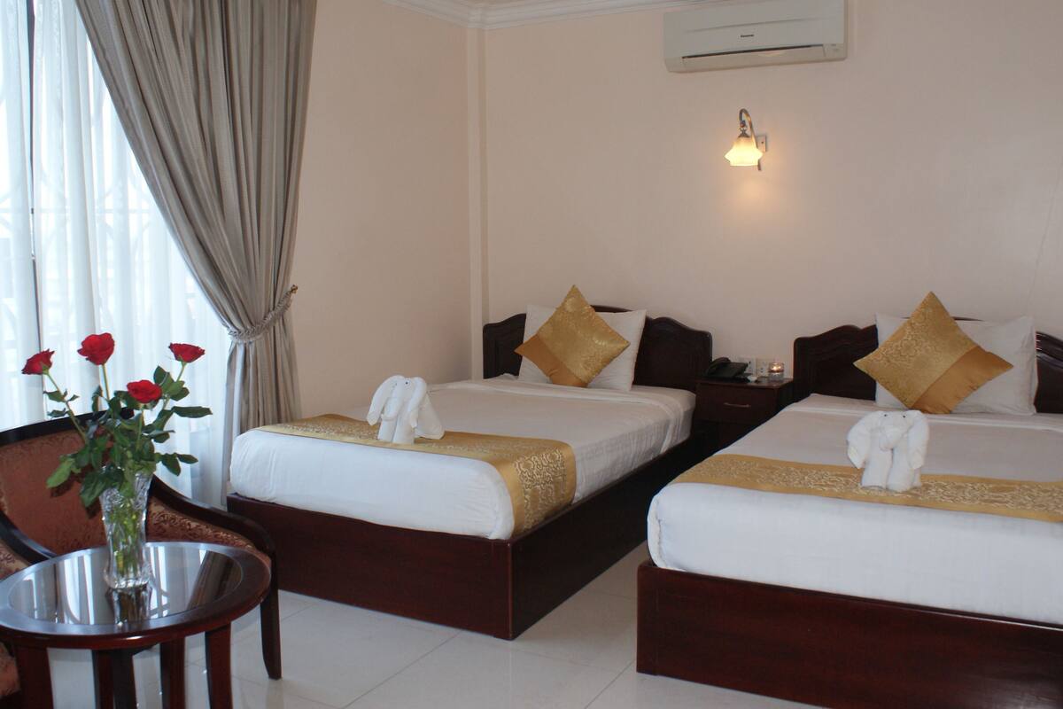 A hotel room with two beds, a nightstand, a table with a vase of flowers, and a window with curtains.