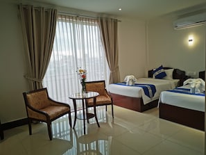 A hotel room with two beds, a small table, two chairs, a vase with flowers, and an air conditioning unit. - Jun Yue Hotel (Phnom Penh)