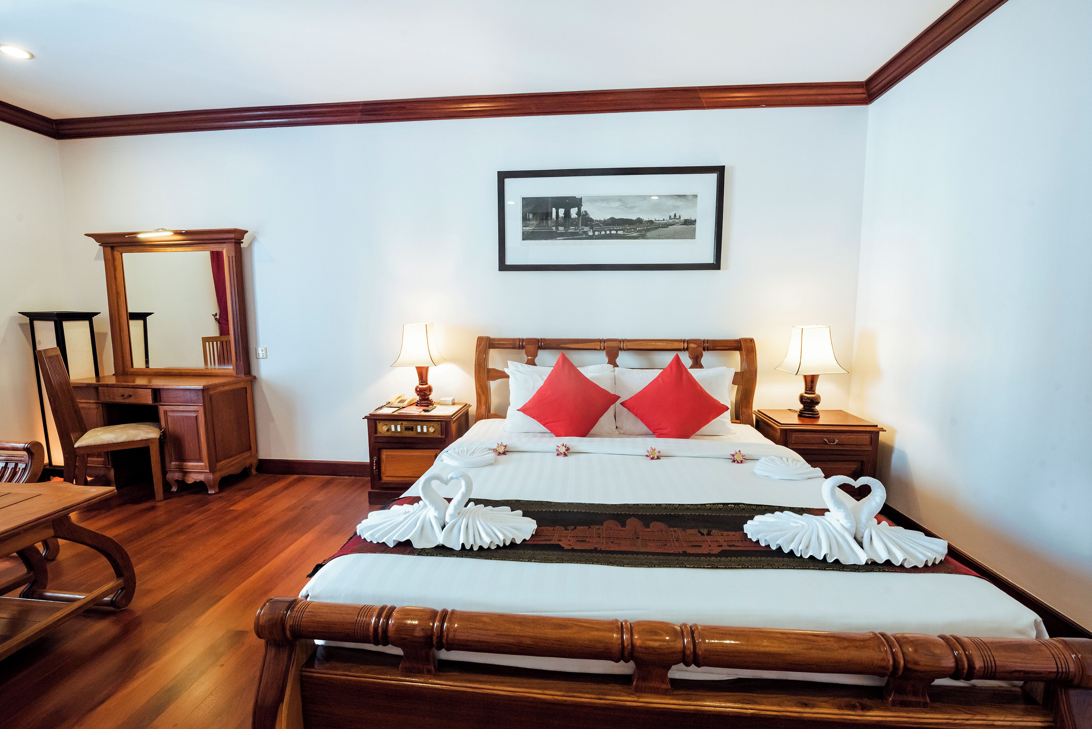A hotel room with a wooden bed, two bedside tables with lamps, a desk with a chair, and a large mirror.