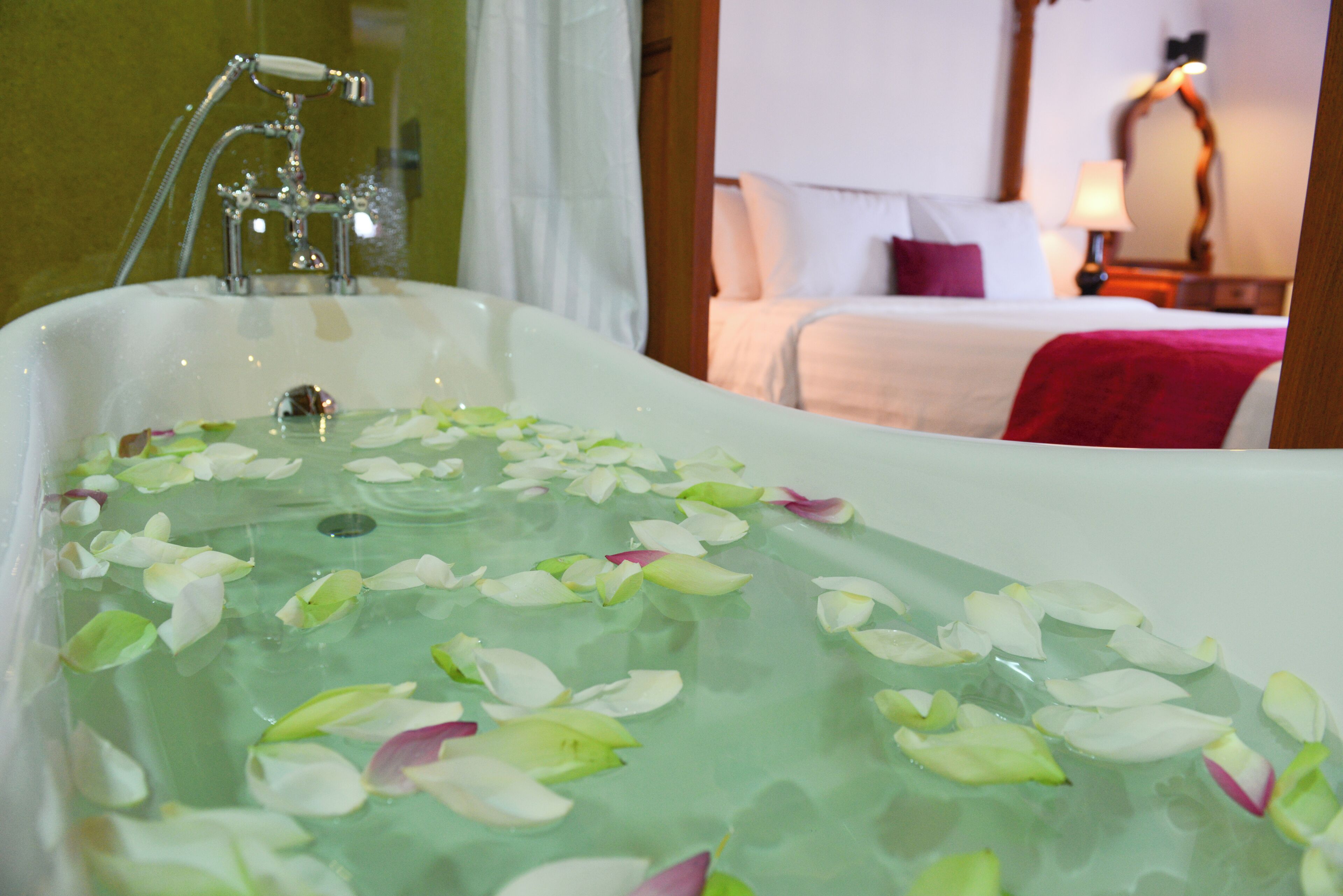 Une baignoire remplie d’eau et de pétales de rose, une pomme de douche et un lit avec une couverture rouge en arrière-plan.