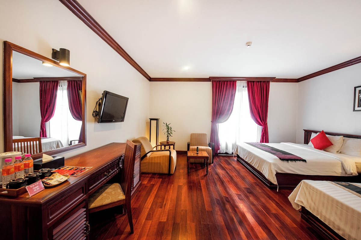 Quarto de hotel com piso de madeira, uma escrivaninha com espelho, televisão, sofá, cadeira, cama e mesa de cabeceira.