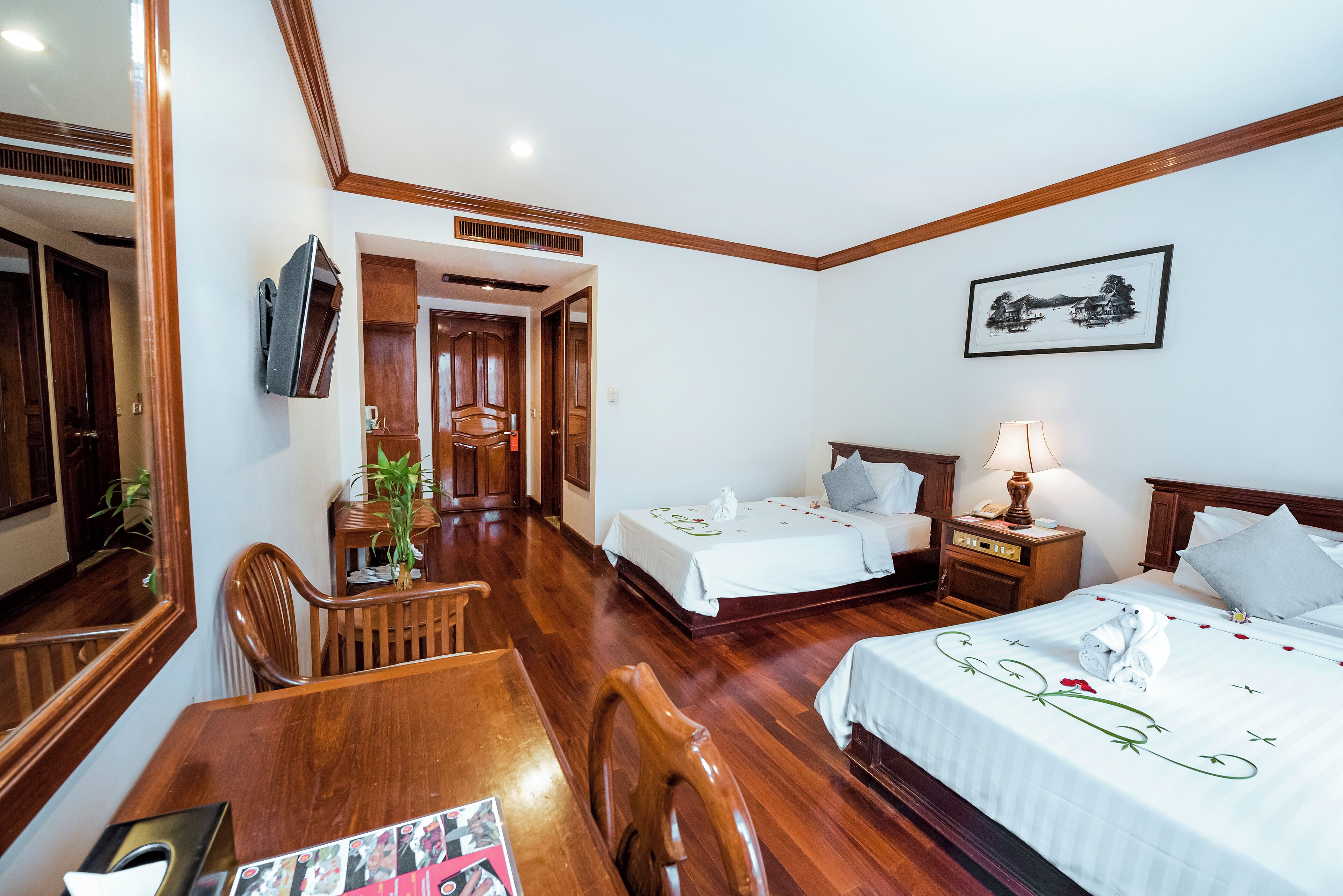 A hotel room with two single beds, a wooden dining table, chairs, a TV mounted on the wall, and a framed picture on the wall.