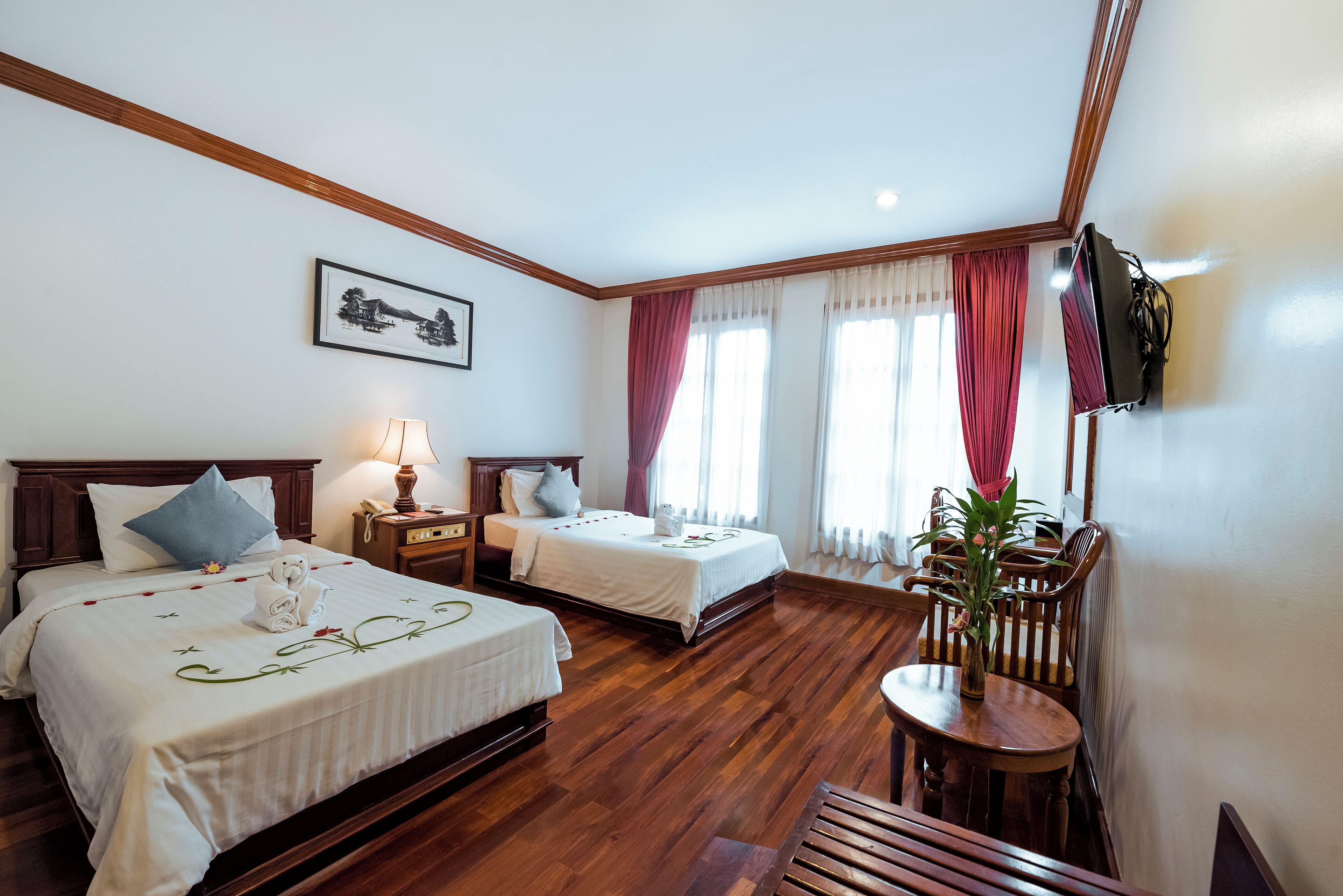 A hotel room with two beds, a wooden floor, a dining area with a table and chairs, a TV mounted on the wall, and a painting above the beds.