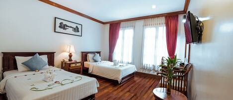 A hotel room with two beds, a wooden floor, a dining area with a table and chairs, a TV mounted on the wall, and a painting above the beds.