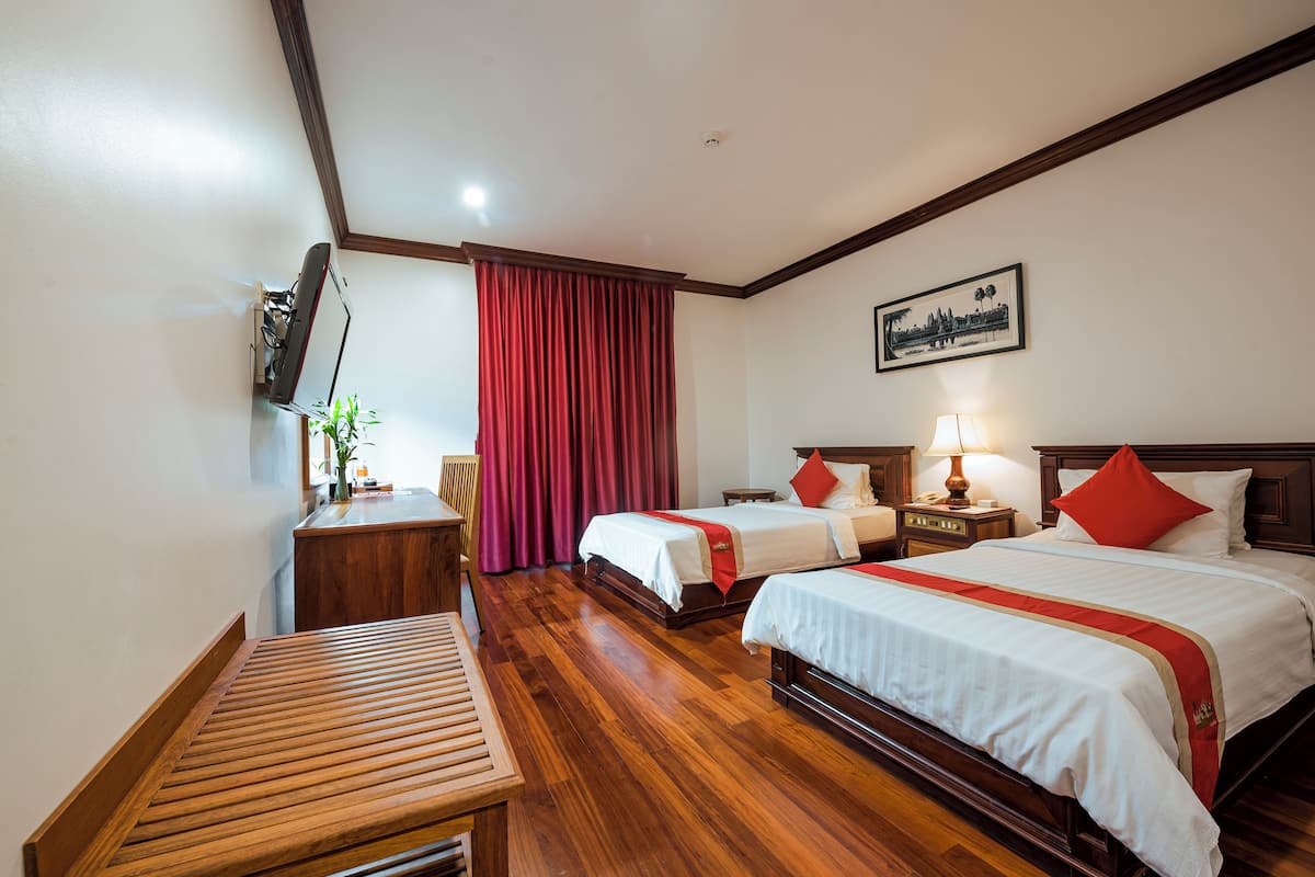 Quarto de hotel com duas camas, um banco de madeira, uma escrivaninha, uma televisão e uma cortina vermelha.