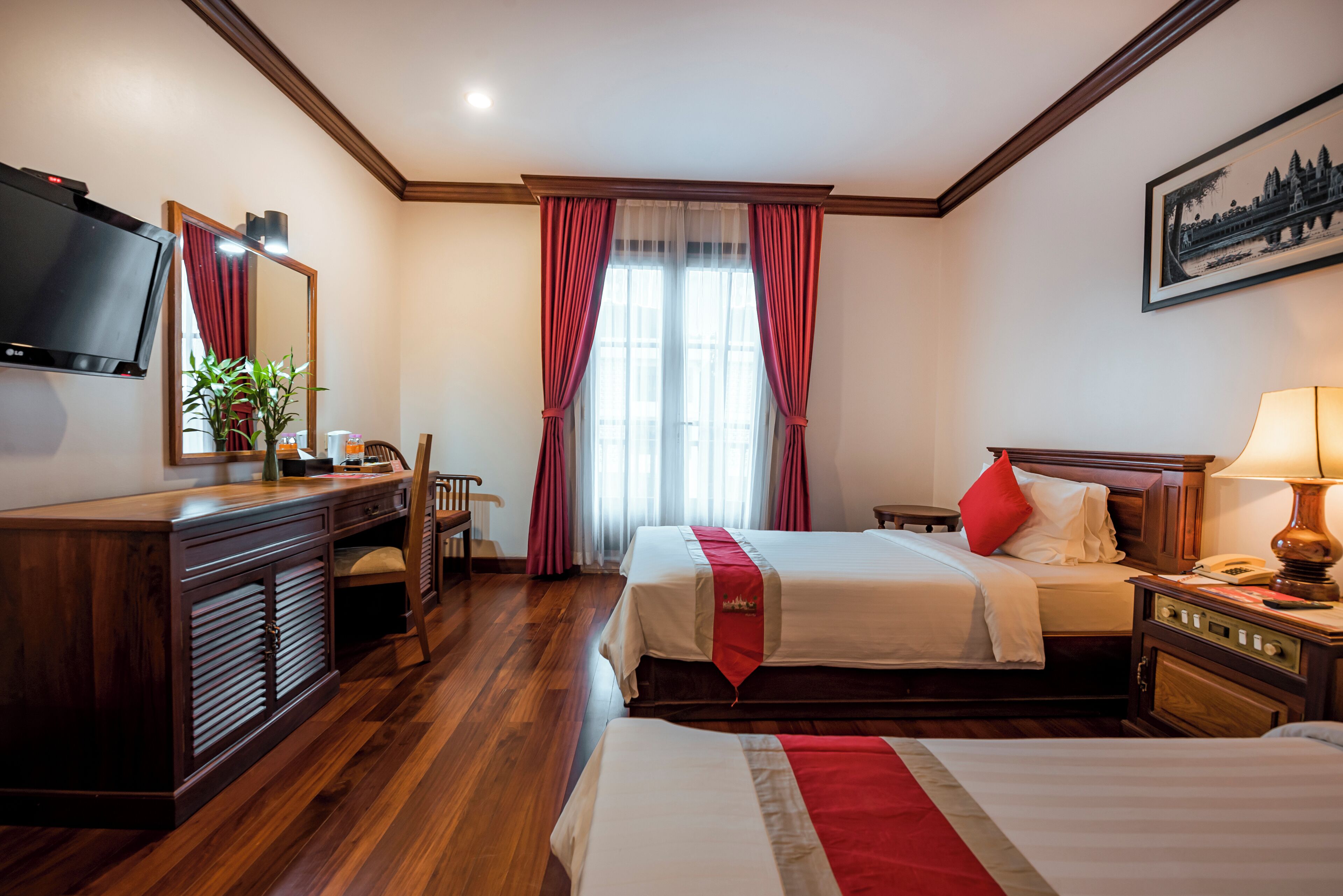 Habitación de hotel con escritorio de madera, televisor, cama con edredón rojo y blanco, y ventana con cortinas rojas.