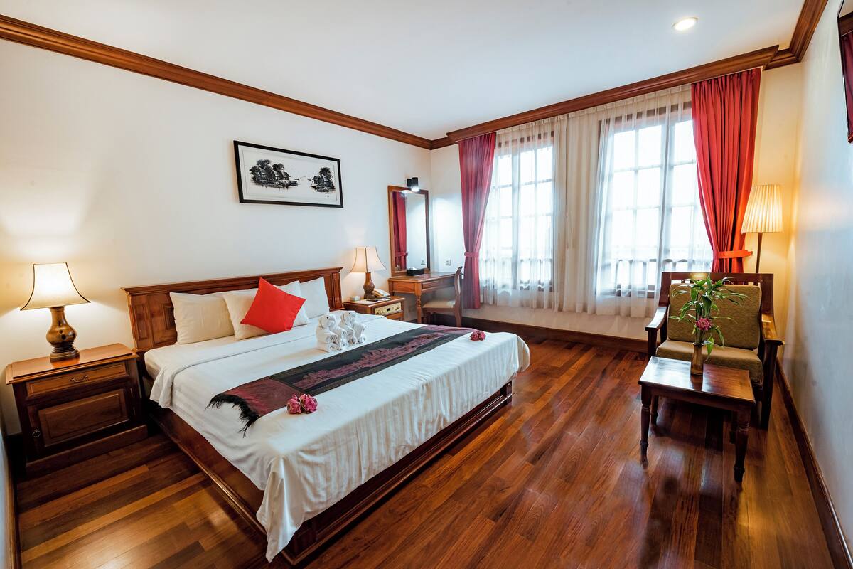 Quarto de hotel com uma cama grande, piso de madeira, mesa de cabeceira com luminária, cadeira com um vaso e janela com cortinas.