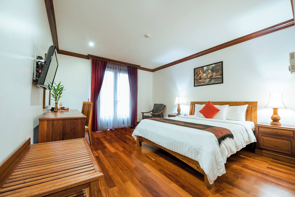Quarto de hotel com cama de madeira, uma escrivaninha, uma cadeira, televisão na parede e janela com cortinas.