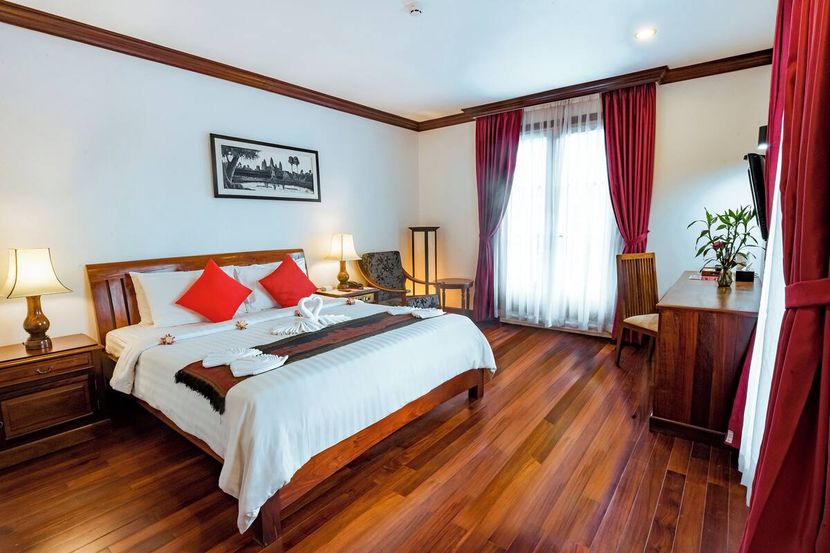 Quarto de hotel com cama de madeira, almofadas vermelhas, mesa de cabeceira com abajur, uma cadeira, escrivaninha e televisão.