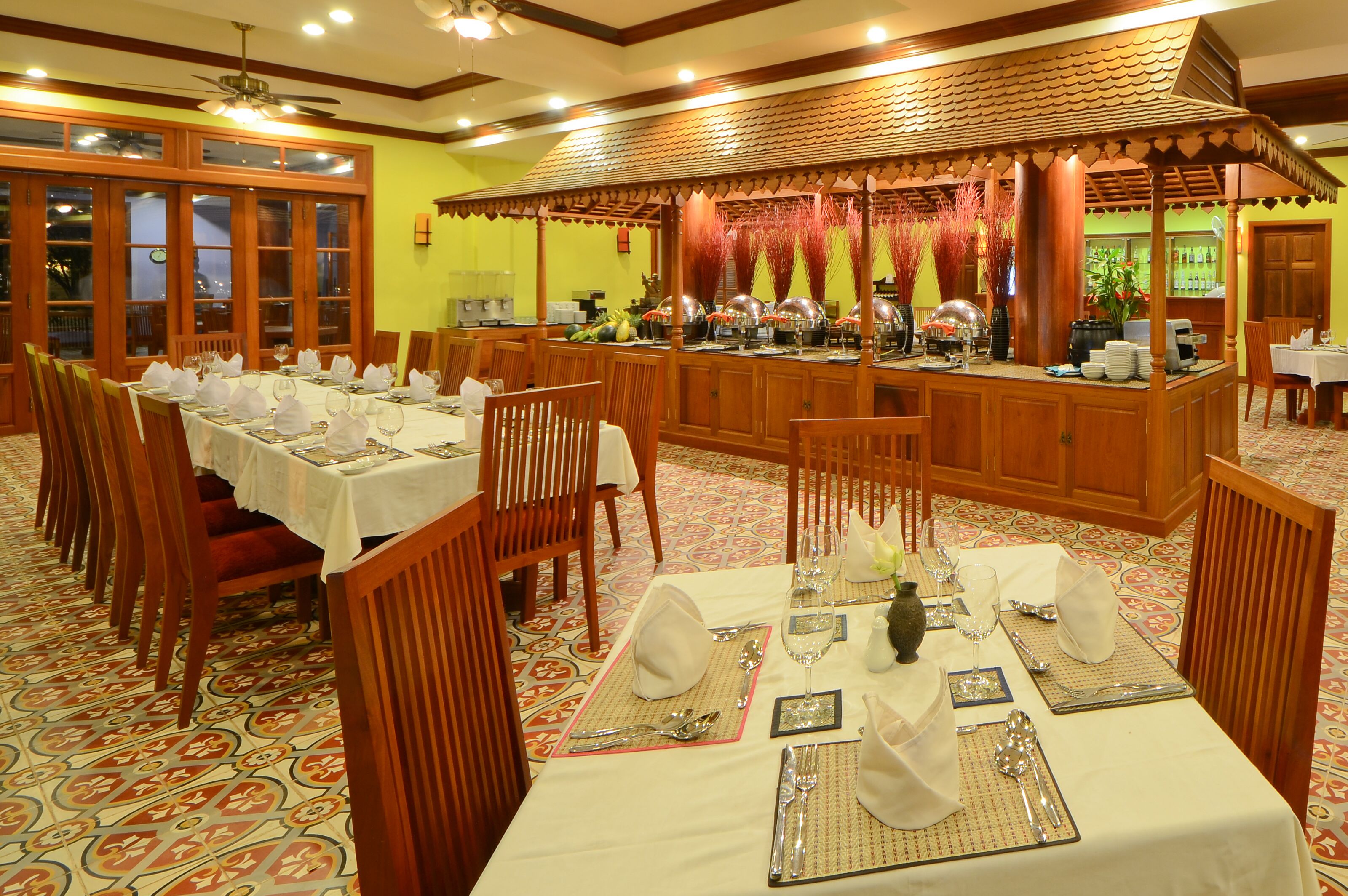 Een restaurant met meerdere tafels gedekt voor diners, houten stoelen en een buffet met diverse gerechten.