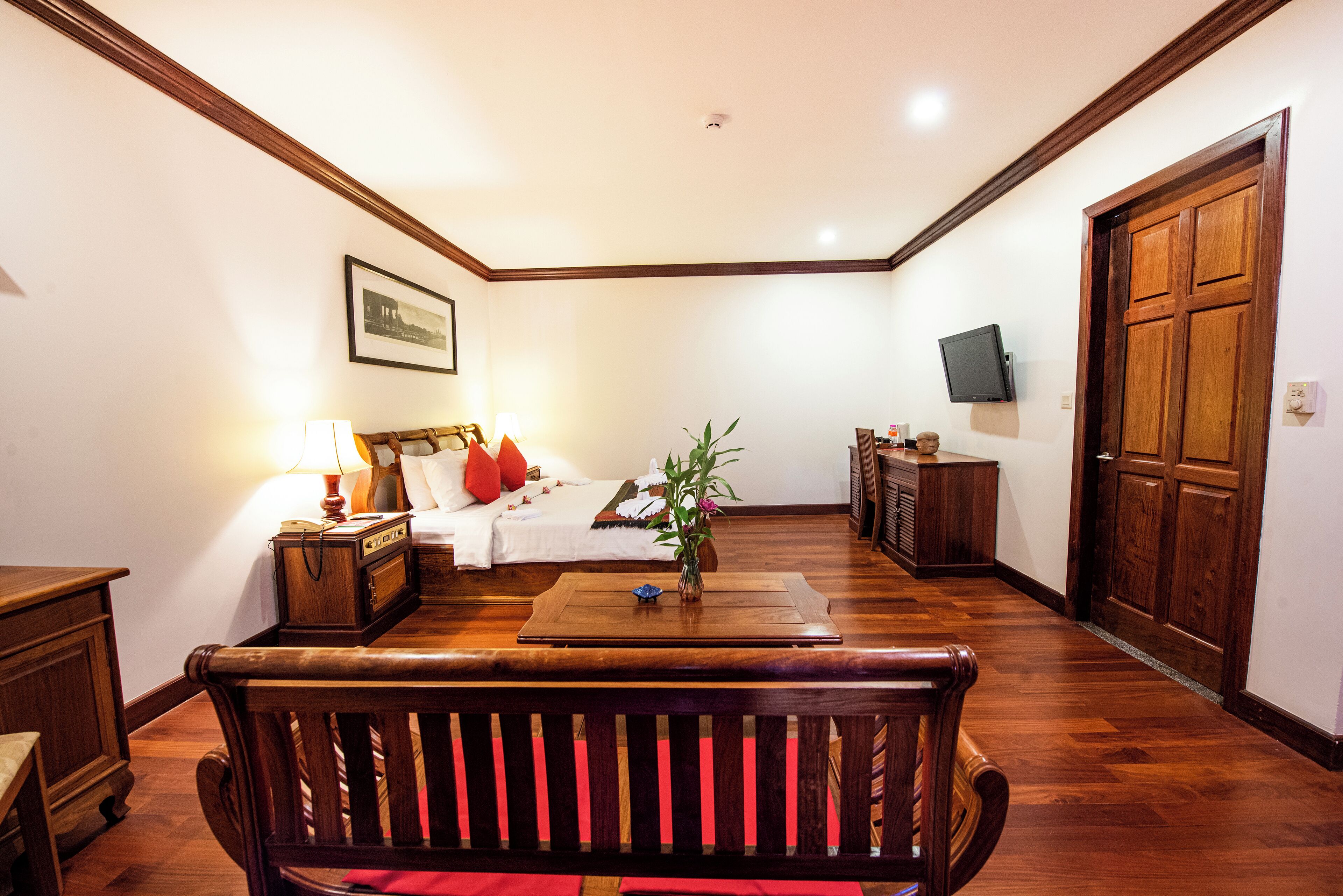 Une chambre d’hôtel avec un plancher en bois, un lit avec des oreillers rouges, une table de chevet avec une lampe, un téléviseur fixé au mur et une porte en bois.