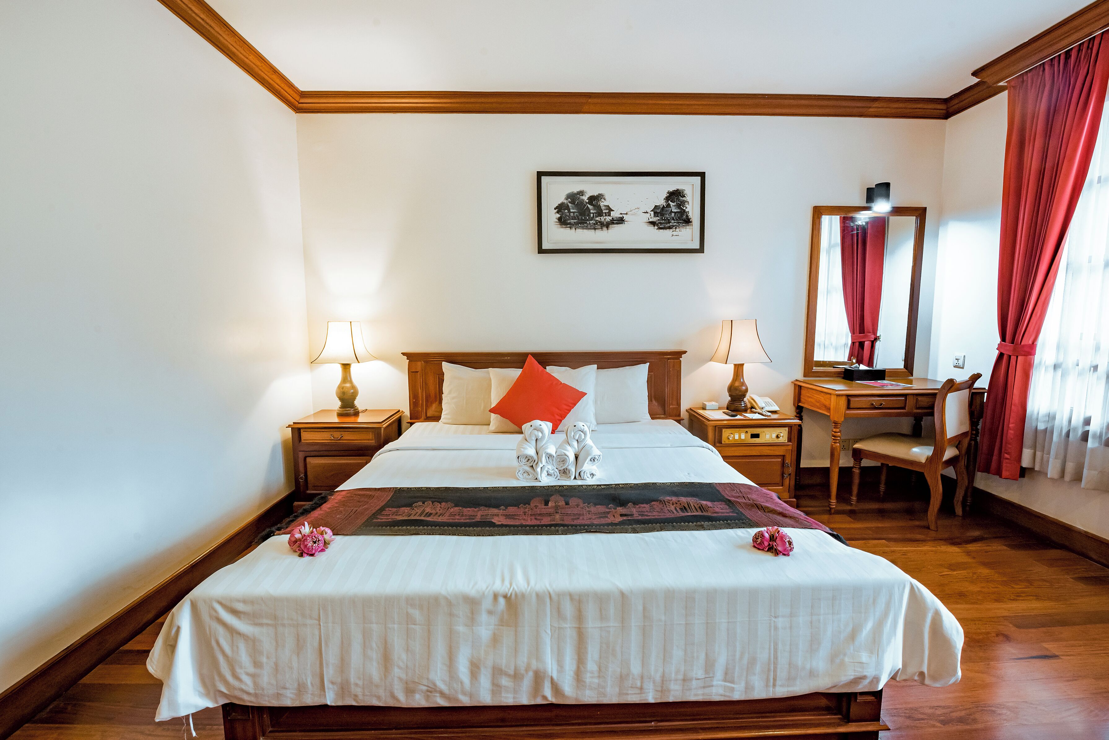 Una cama bien hecha con un almohadón rojo, dos mesitas de noche con lámparas, un escritorio de madera con una silla y un cuadro en la pared.
