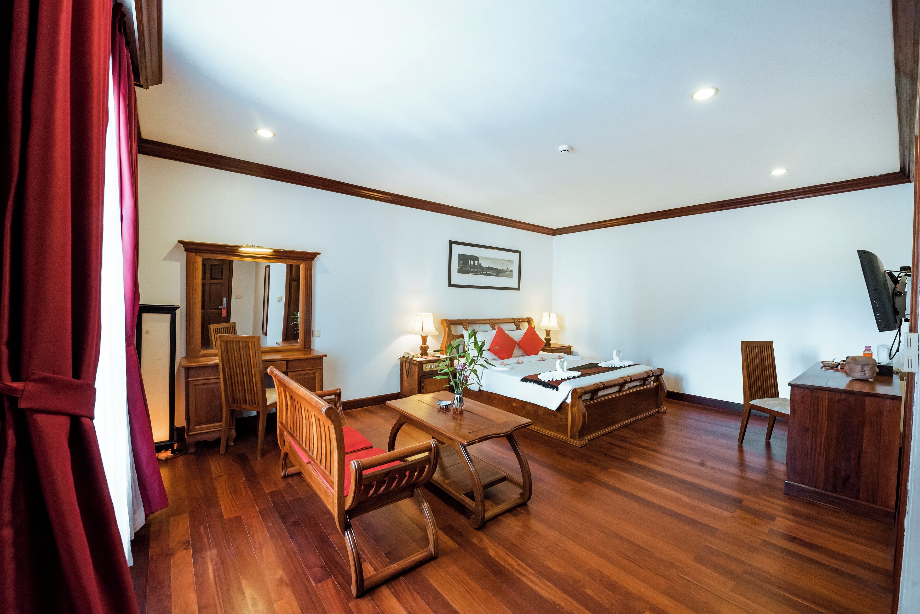 A hotel room with a wooden floor, a bed with red pillows, a desk with a chair, a wooden cabinet, and a television on a stand.