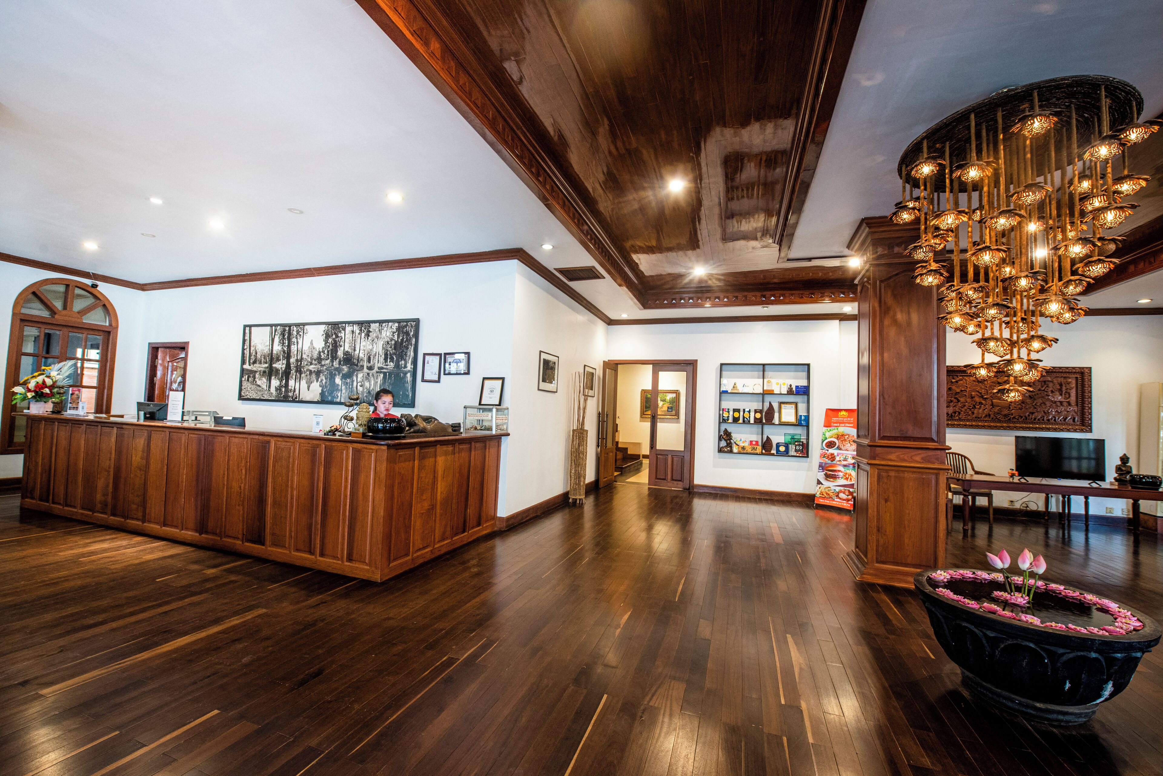 A spacious lobby with a wooden reception desk, a large chandelier, and a reception area with framed pictures and a television.
