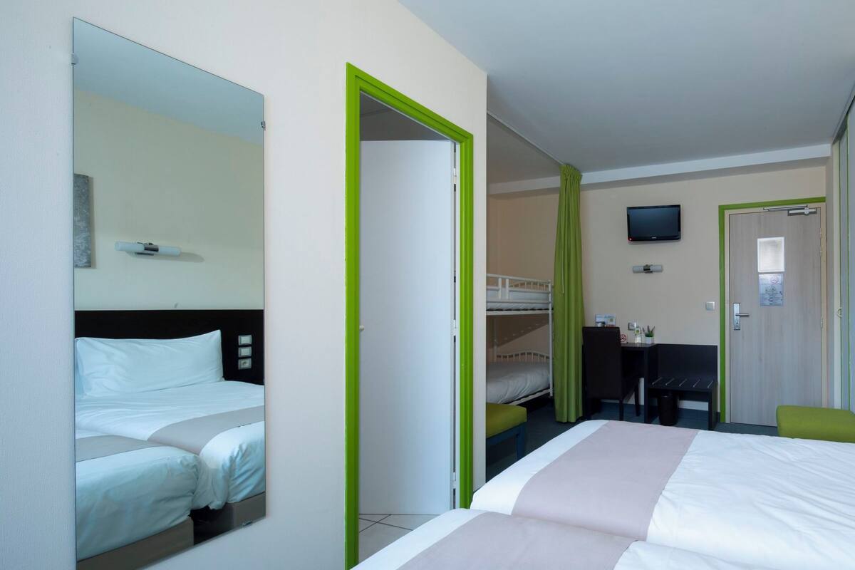 Quarto de hotel com cama, escrivaninha, cadeira e televisão na parede.