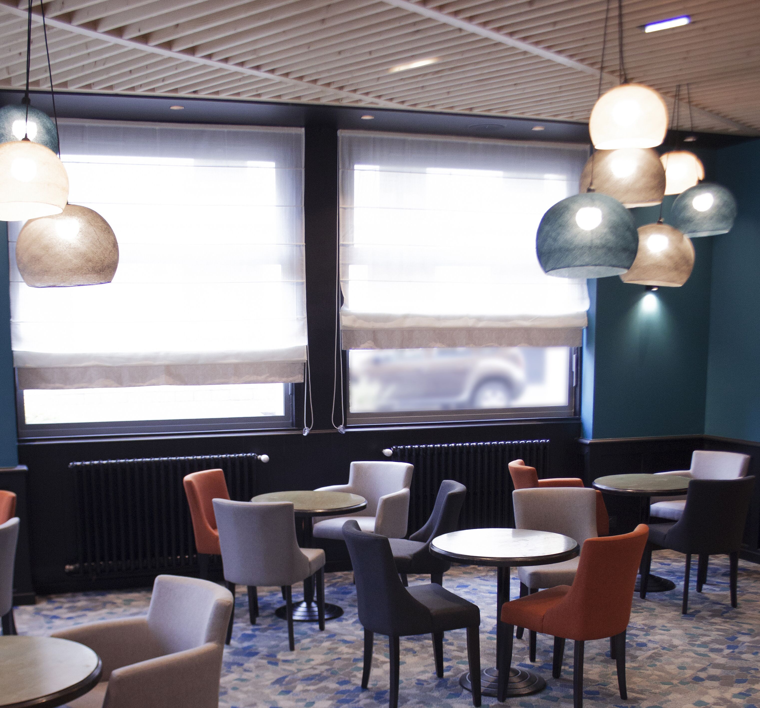 Un restaurante con mesas y sillas redondas, ventanas amplias con persianas y lámparas colgantes.