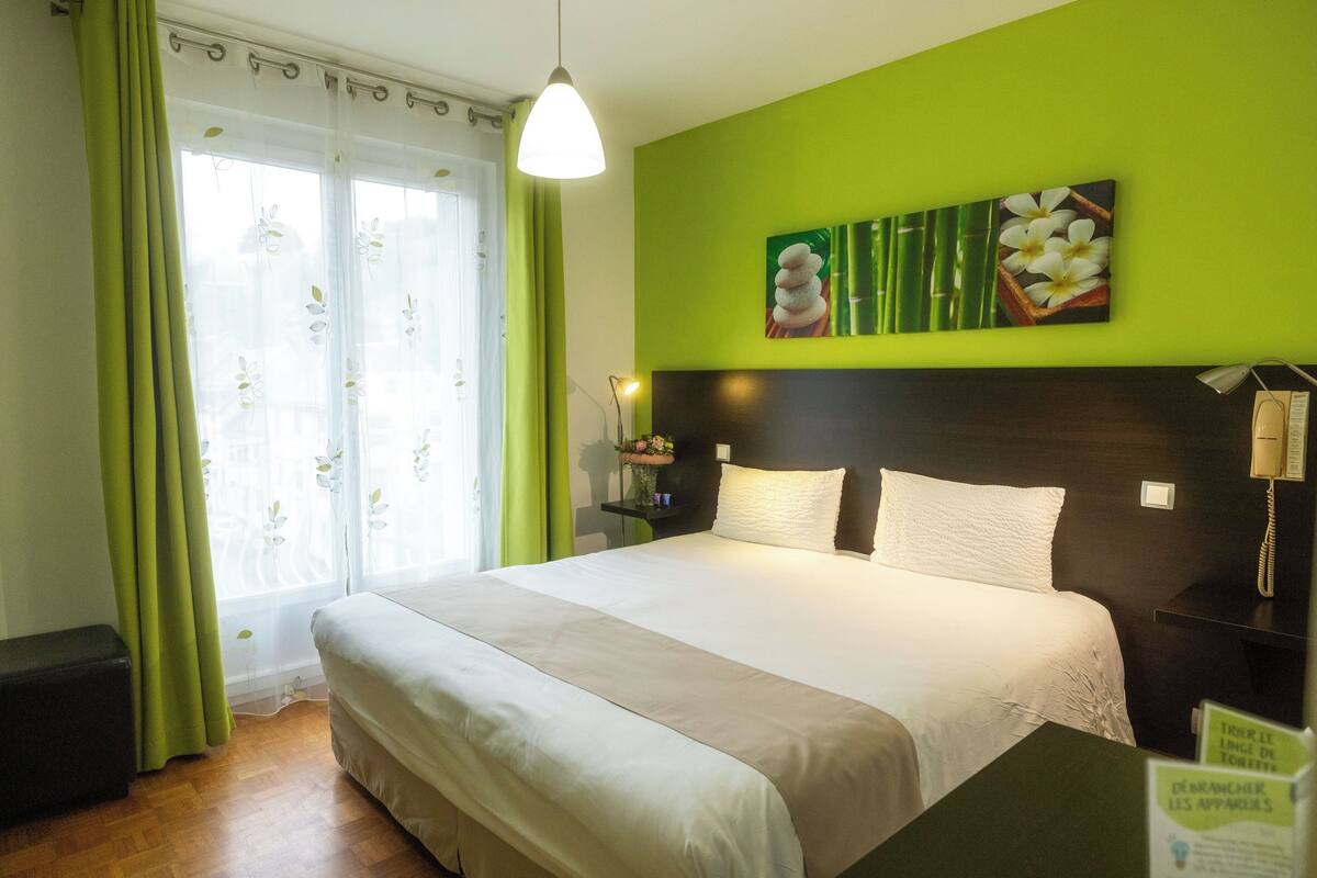 Quarto de hotel com uma cama grande, uma mesinha de cabeceira com abajur, uma parede verde e uma janela com cortinas finas.