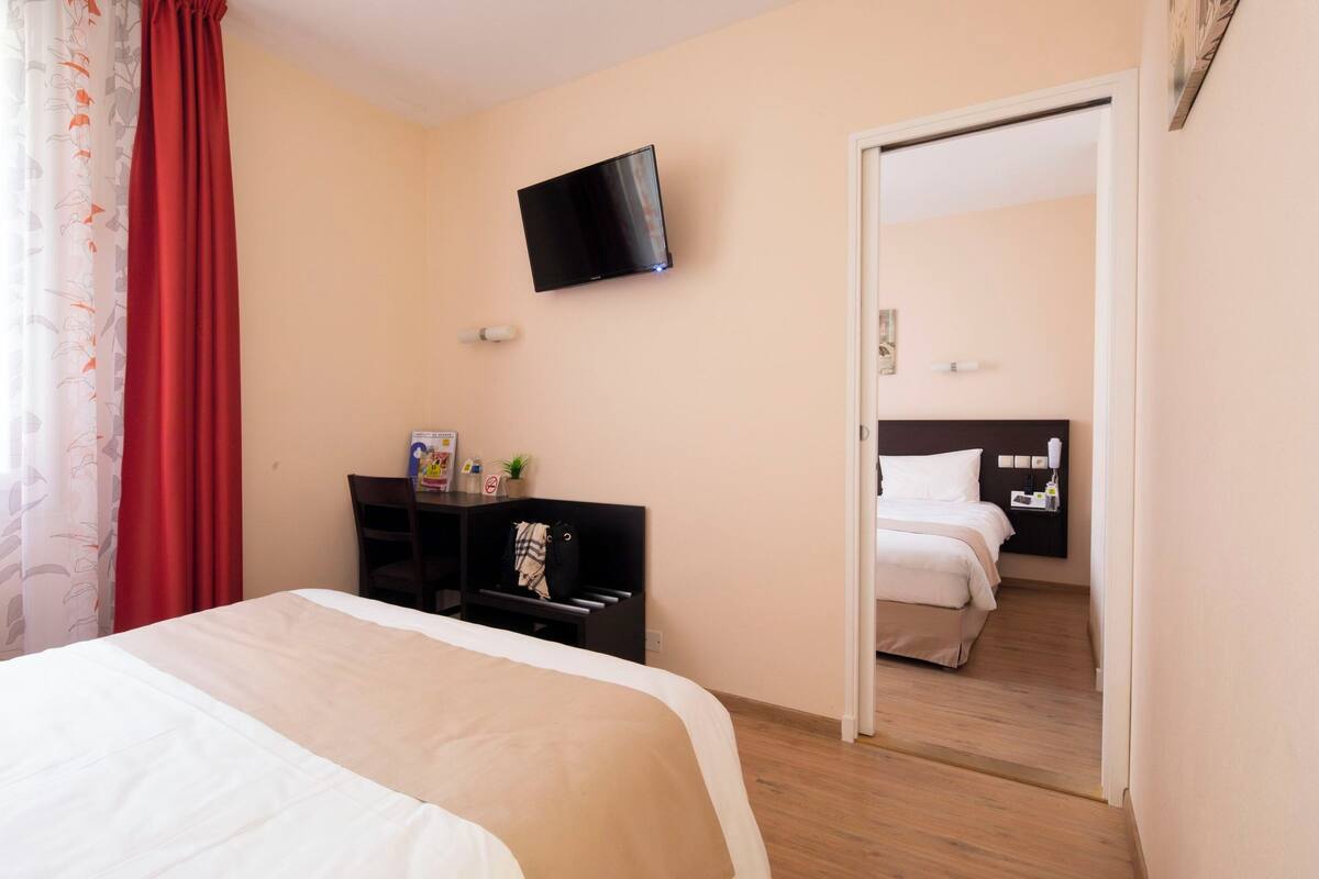 Quarto de hotel com cama, escrivaninha com cadeira, televisão fixada na parede e porta que dá para outro cômodo.