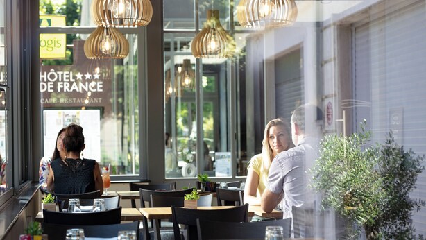 Une vue intérieure d’un restaurant, avec des tables et des chaises, une plante en pot et une vue sur l’extérieur.