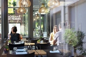 Ein Blick ins Restaurantinnere zeigt Tische und Stühle, einen Blumentopf und einen Ausblick nach draußen.