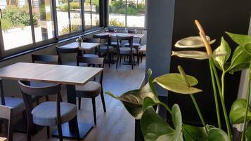 A restaurant interior with wooden tables and chairs, large windows, and indoor plants.