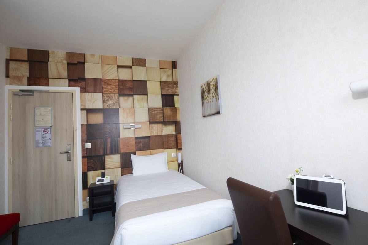 Quarto de hotel com cama, mesa de cabeceira, cadeira, televisão e uma parede com detalhe em painel de madeira.