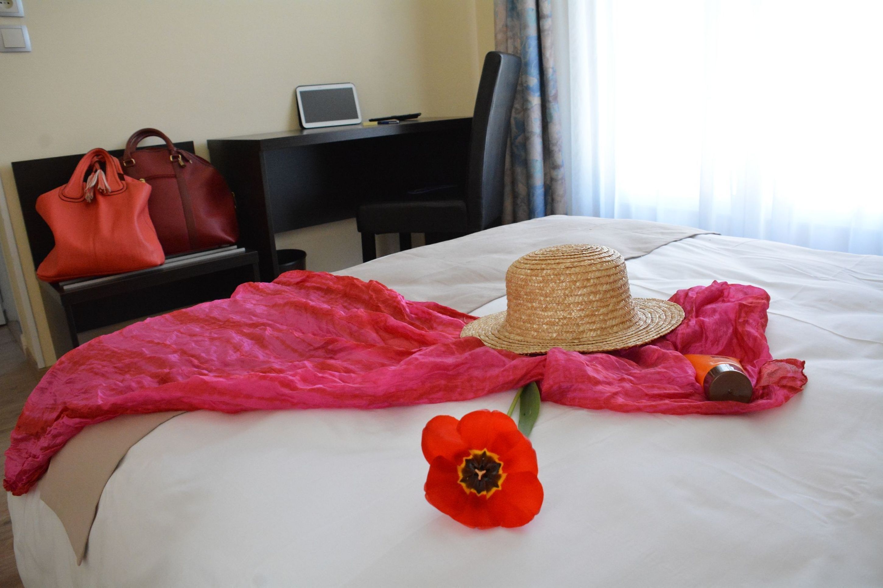 Habitación de hotel con cama, una laptop en un escritorio, un bolso rojo, un sombrero de paja, una flor roja y una vela.