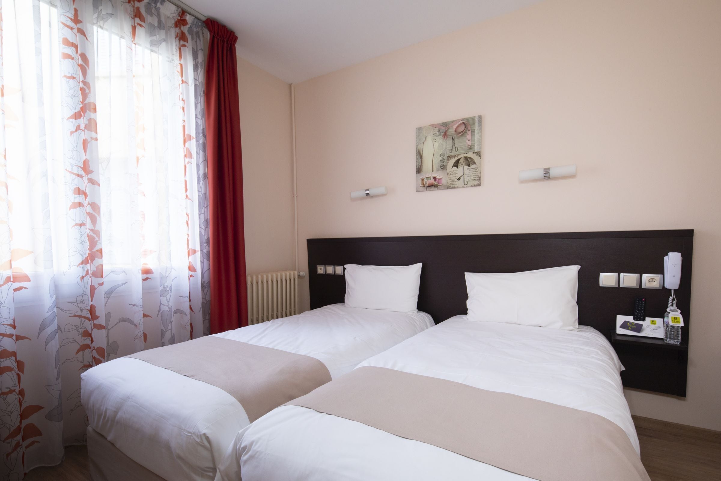 A hotel room with two beds, a window with curtains, a wall-mounted artwork, and a bedside table with a phone and a bottle.