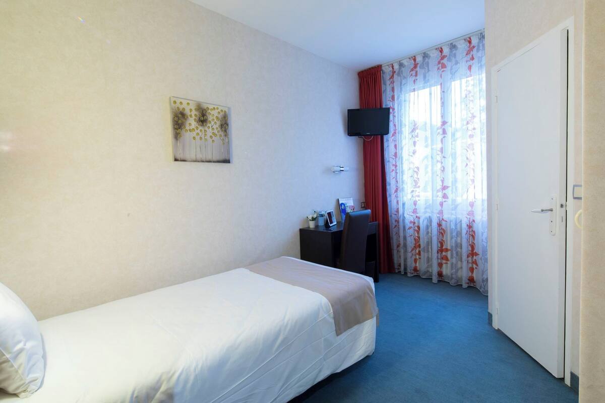 Quarto de hotel com uma cama de solteiro, uma mesa com cadeira, televisão e uma porta.