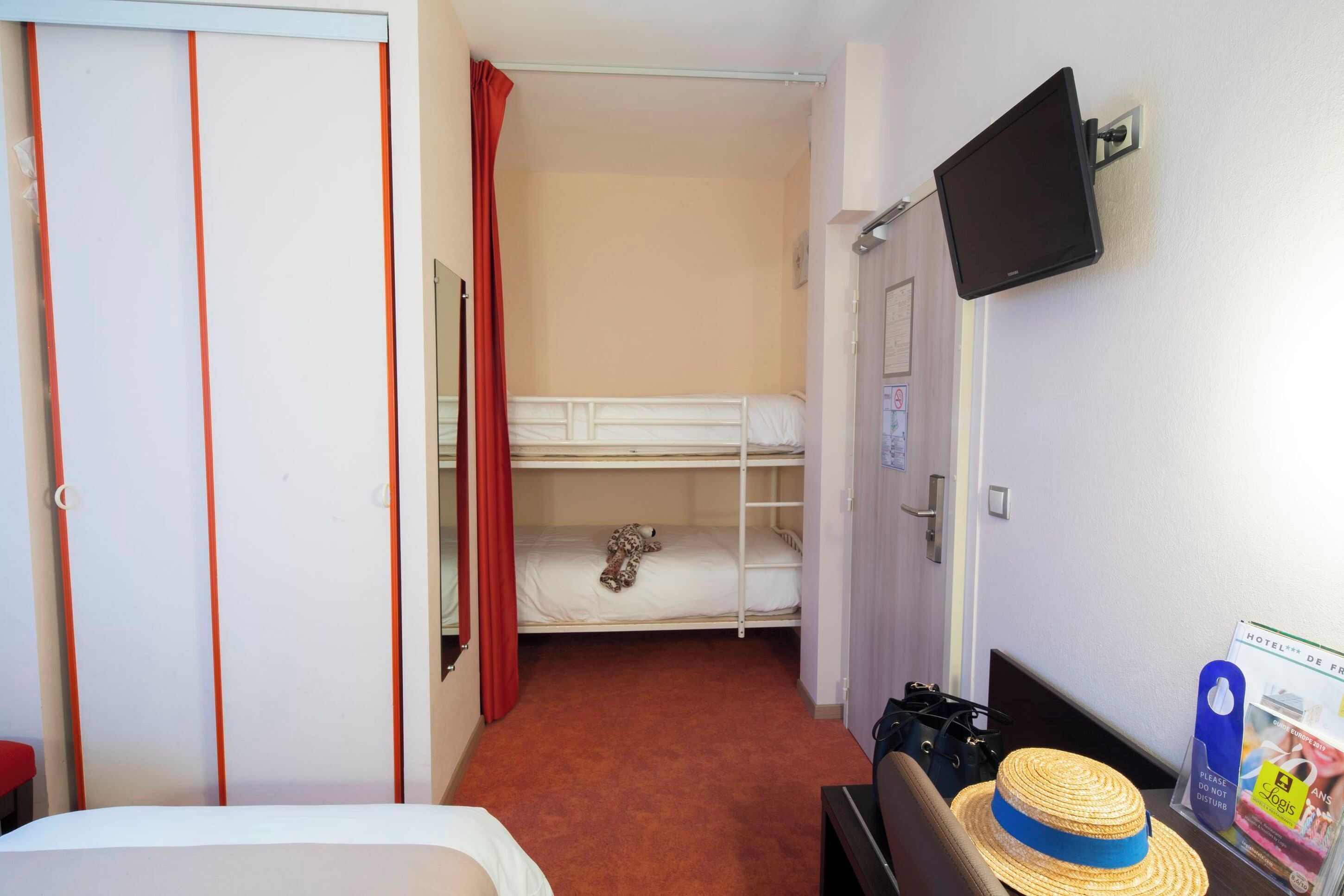 Habitación de hotel con literas, cama plana, televisor montado en la pared, escritorio con silla y un sombrero sobre el escritorio, además de una cortina roja.