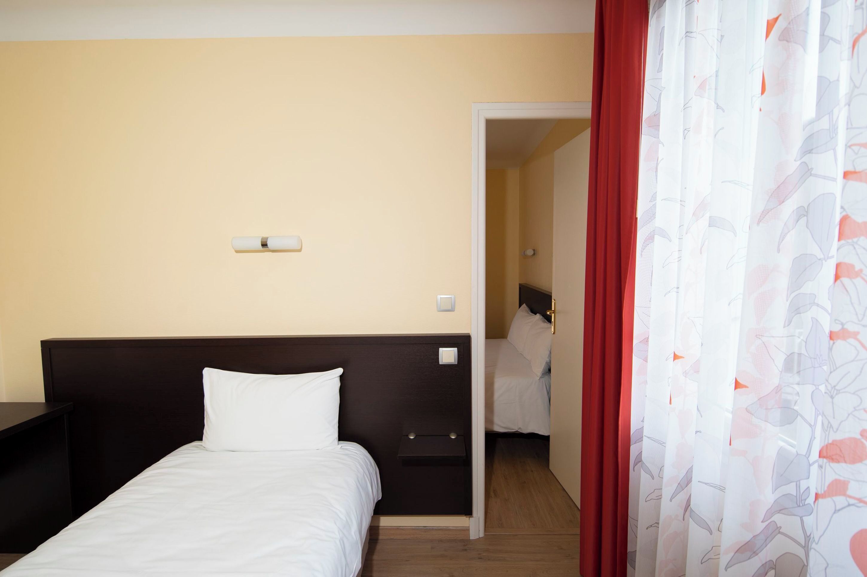 A hotel room with a bed, a nightstand, a door leading to another room, and a curtain with a floral pattern.