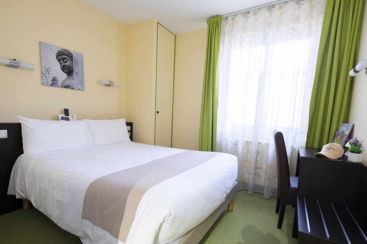 Cama bem arrumada com lençóis brancos, mesa de cabeceira com abajur, uma cadeira e escrivaninha com um boné e uma pequena planta.