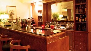 Una zona de bar con mostrador de madera, taburetes y estantes con botellas y vasos.