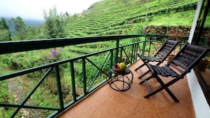A balcony with two wooden lounge chairs, a small round table with a fruit basket, and a view of terraced tea plantations. - Camelot Resort (Devikolam)