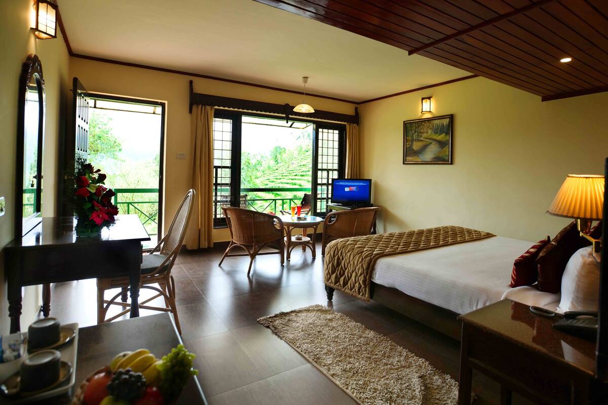 Une chambre d’hôtel comprenant un lit, une télévision, une table à manger avec des chaises et un balcon donnant sur un espace verdoyant.