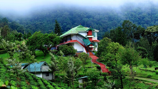 A house with a green roof and red accents, surrounded by lush greenery and a misty forest backdrop. - Camelot Resort (Devikolam)
