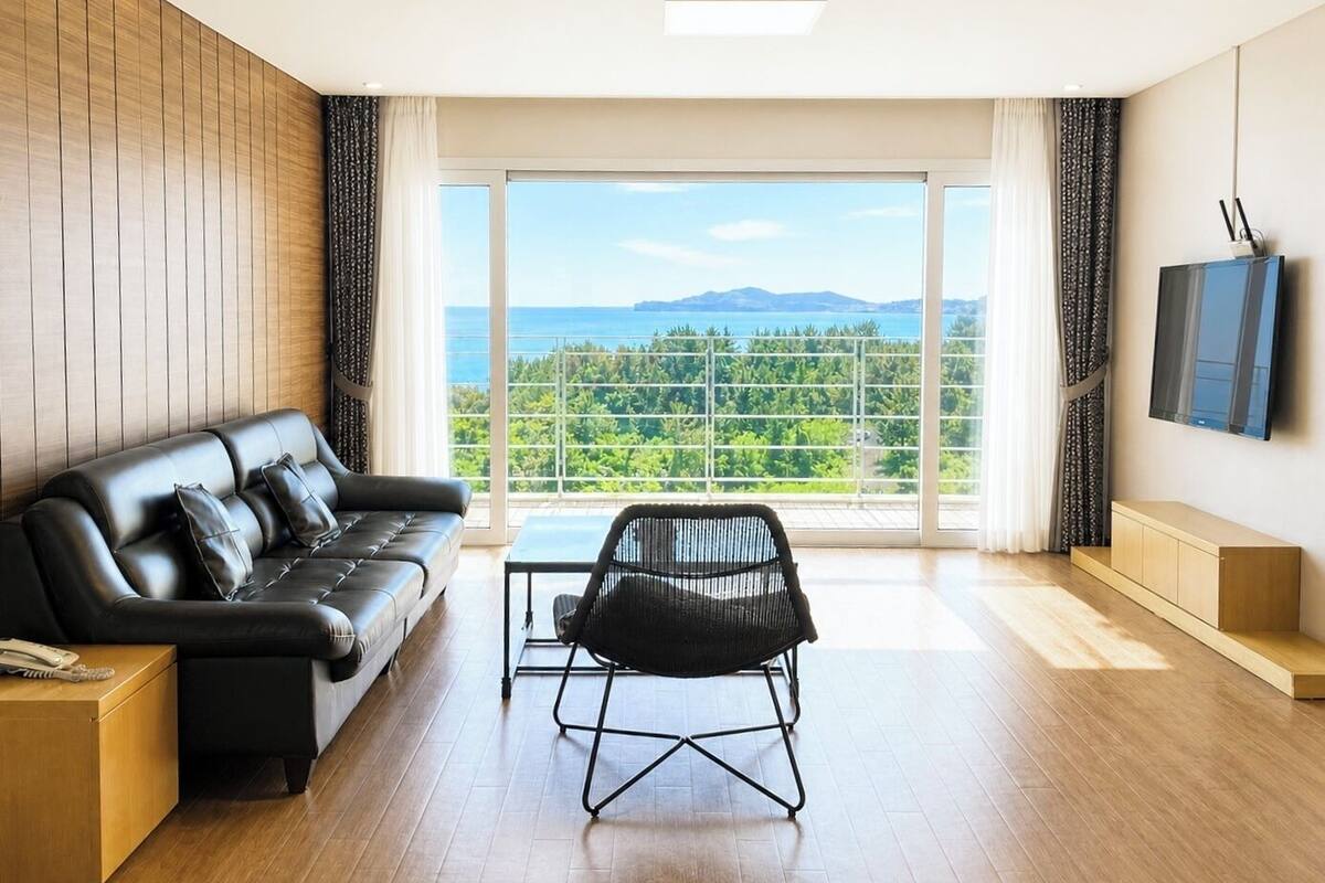 A modern living room with a leather sofa, a black chair, a TV, and a large window with a view of greenery and mountains.