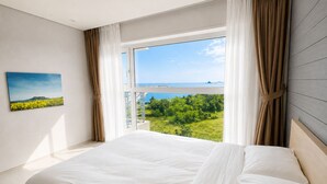 A bedroom with a large bed, a window with curtains, a painting on the wall, and a view of greenery and water. - Y Resort Jeju (Seogwipo)