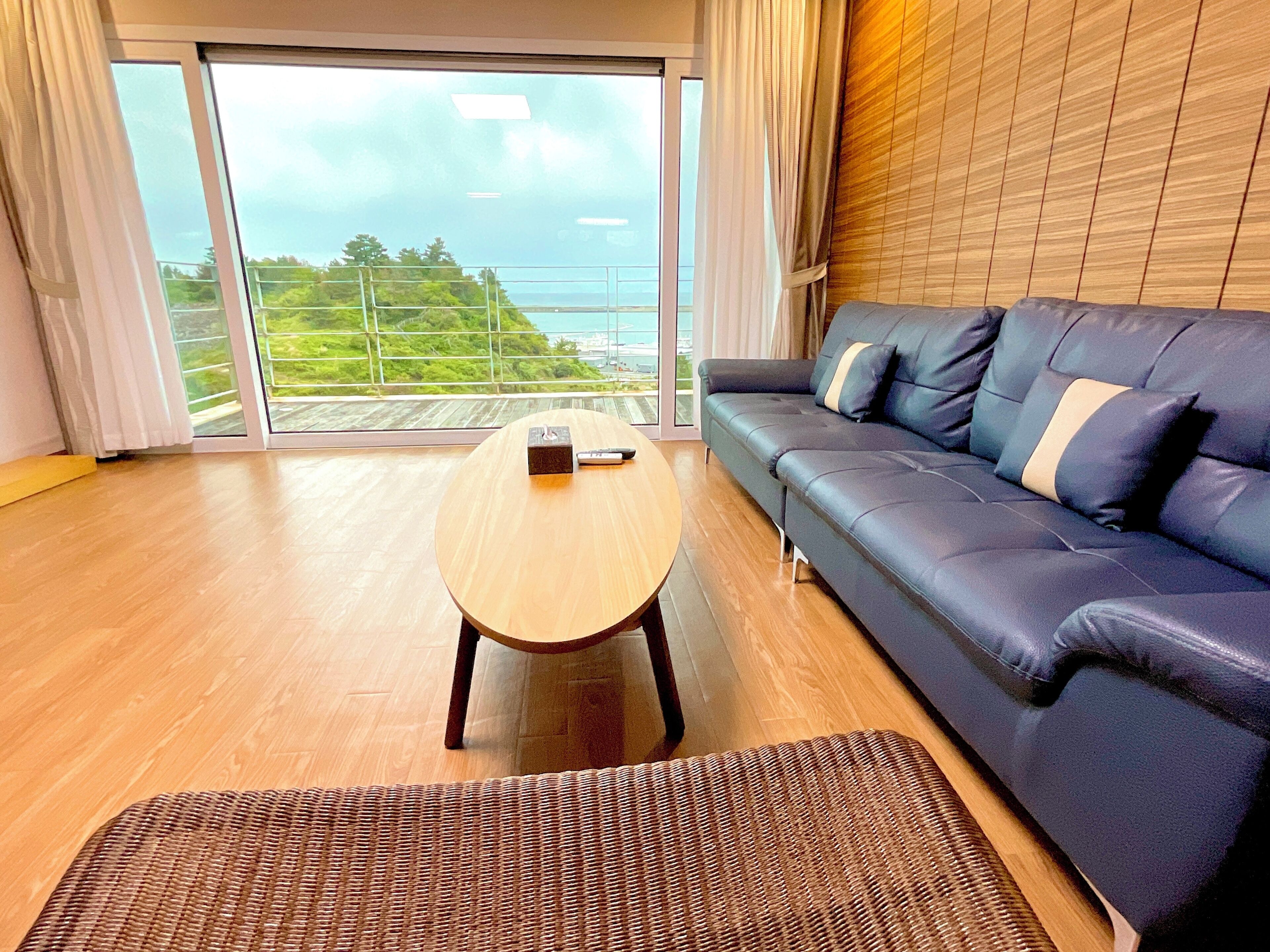 Ein modernes Wohnzimmer mit Holzboden, einem dunkelblauen Sofa, einem runden Holztisch und Blick auf Grünflächen sowie ein Gewässer durch große Glasüren.