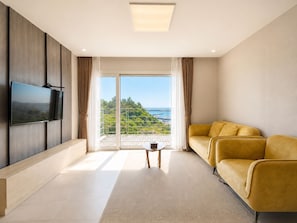 Un salon moderne avec un téléviseur à écran plat, un canapé jaune, une petite table basse et une grande porte-fenêtre coulissante donnant sur un balcon avec vue sur la mer.