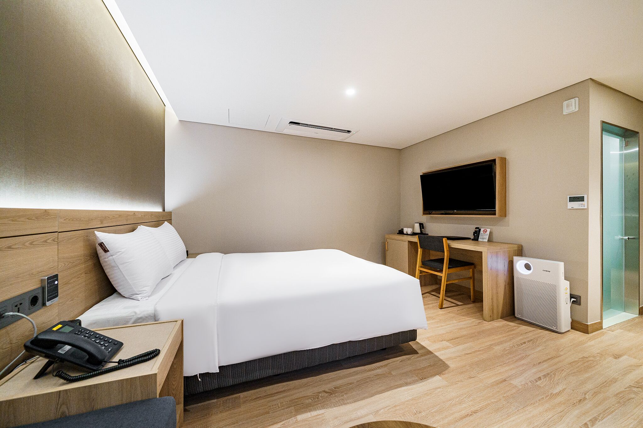 A modern hotel room with a bed, bedside telephone, desk, chair, television, and a wall-mounted air conditioning unit.