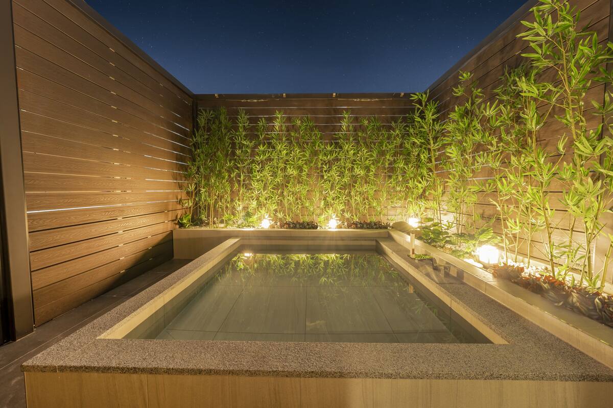 Ein rechteckiges Whirlpool mit einer umlaufenden Holzterrasse, vor einer Kulisse aus Bambuspflanzen und einer Holzwand.