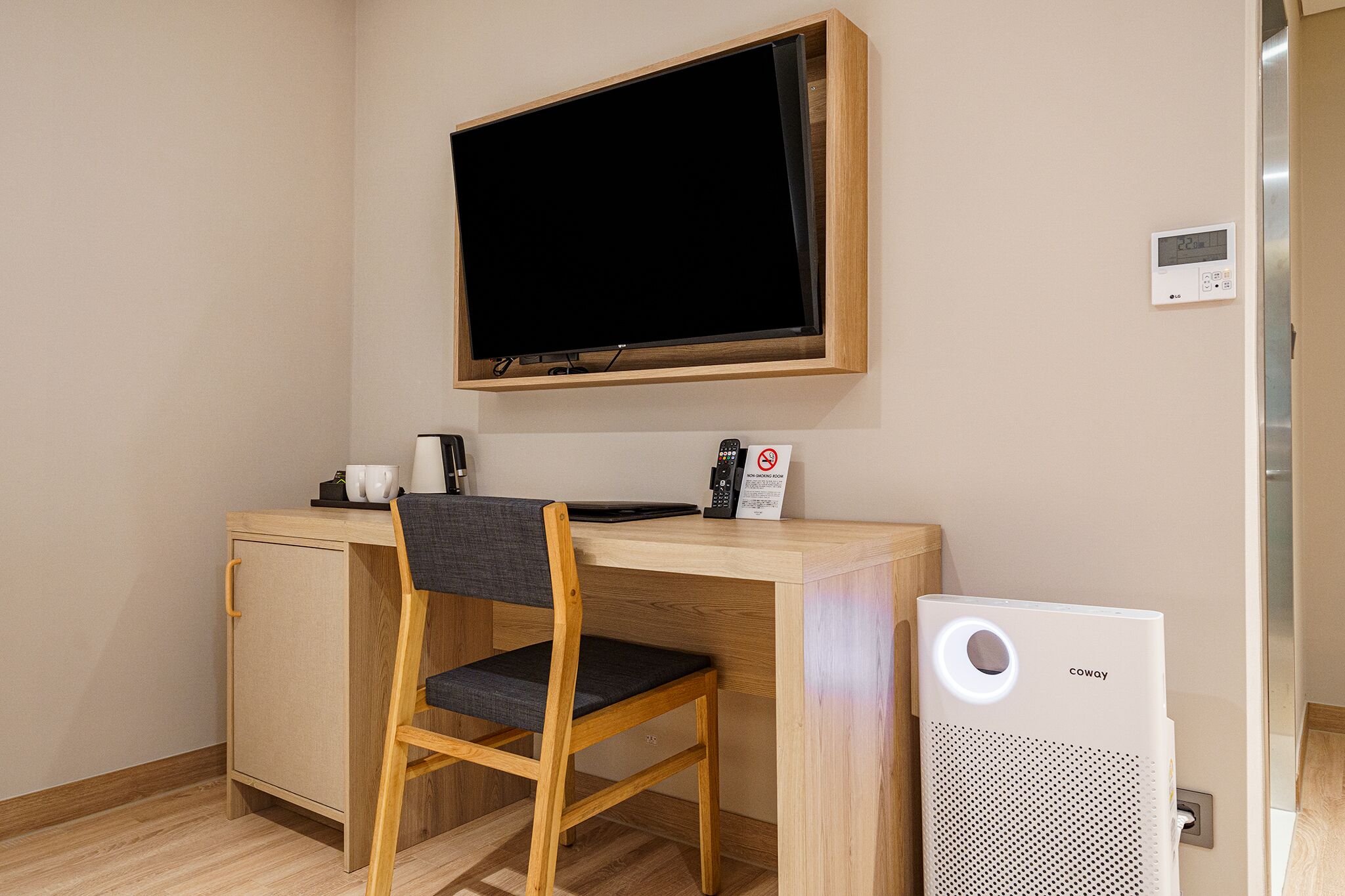 A small, well-lit room with a wooden desk, a chair, a television mounted on the wall, and a white air purifier.