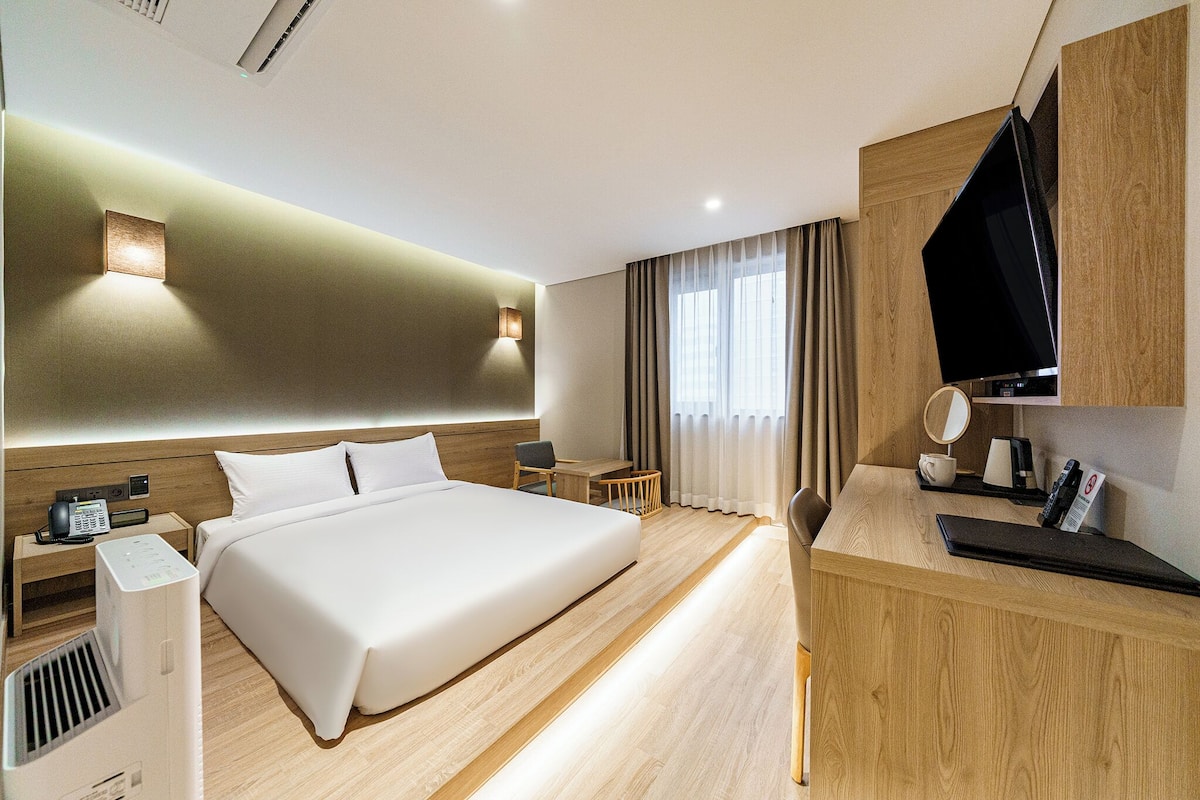 Ein Hotelzimmer mit einem großen Bett, Nachttischen, einem Schreibtisch, einem Flachbildfernseher und einem Fenster mit Vorhängen.