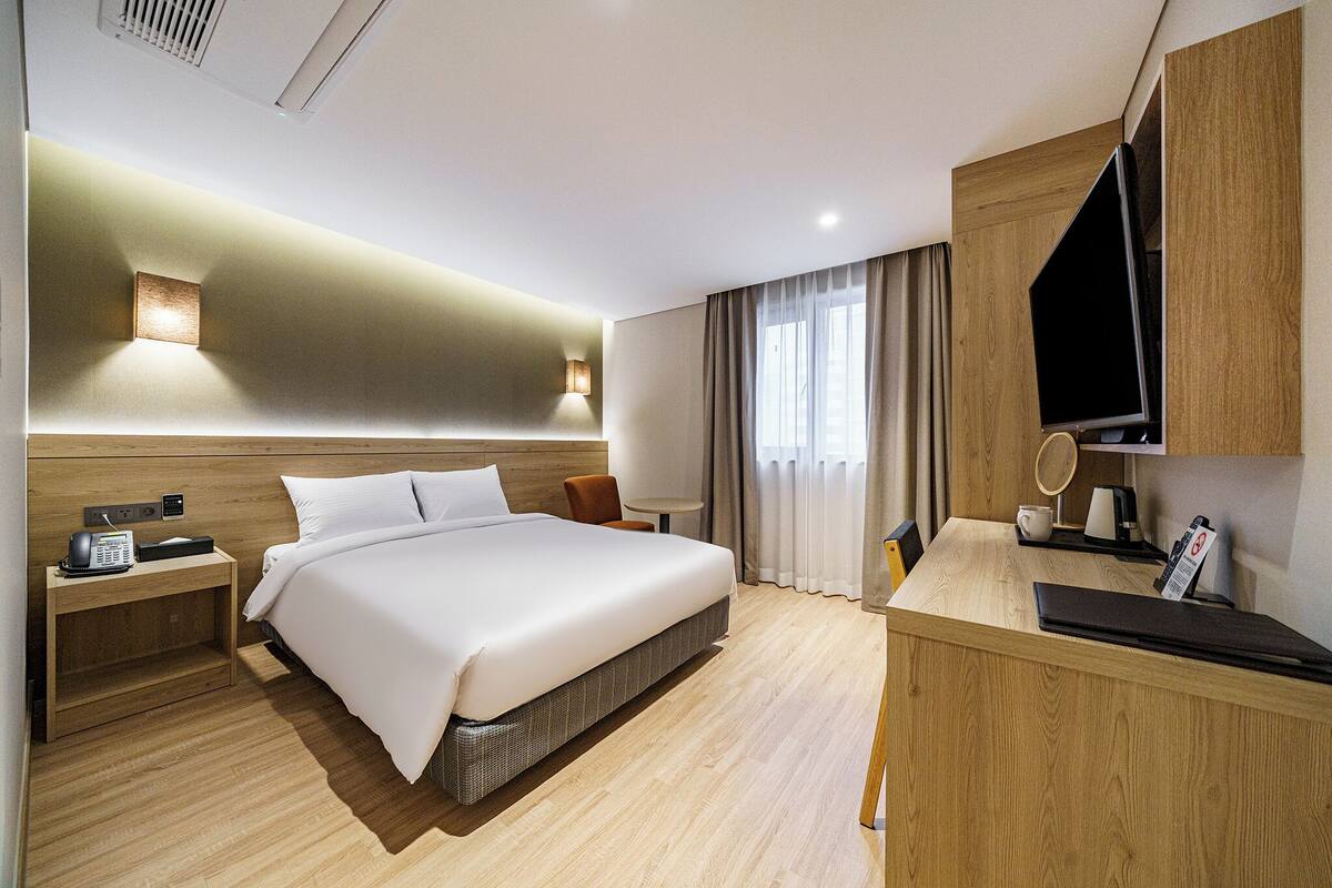 Ein modernes Hotelzimmer mit einem großen Bett, einem Nachttisch mit Telefon, einem Schreibtisch mit Fernseher und einem Fenster mit Vorhängen.