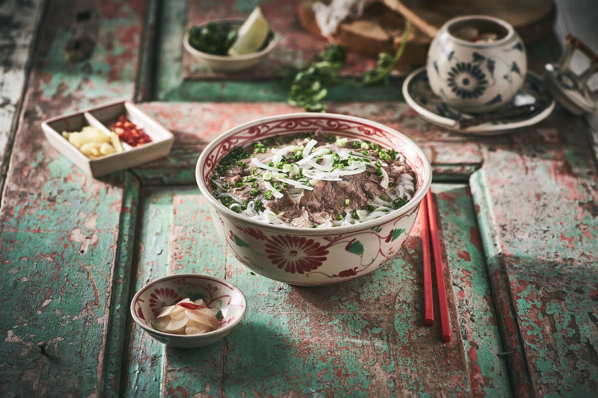 Eine Schüssel Suppe mit Stäbchen und einer kleinen Schale Beilagen auf einem rustikalen Holztisch.