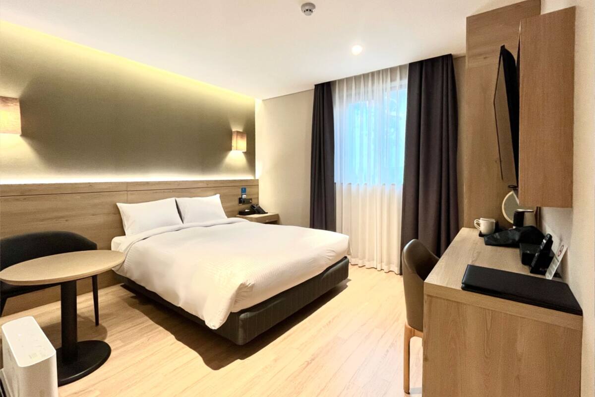 Ein modernes Hotelzimmer mit Bett, Nachttisch, Sessel, Schreibtisch und einem kleinen Tisch mit Tasse und Telefon.